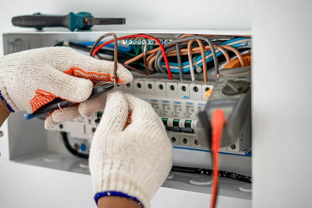 Électricien en gants blancs manipulant des fils colorés dans un tableau électrique. Des disjoncteurs affichent "O-OFF". Un outil et multimètre visibles.