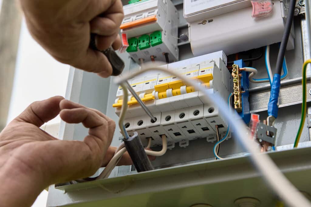 Électricien vissant un disjoncteur dans un tableau électrique. Fils colorés, compteur et indications 230/400V C16 visibles.