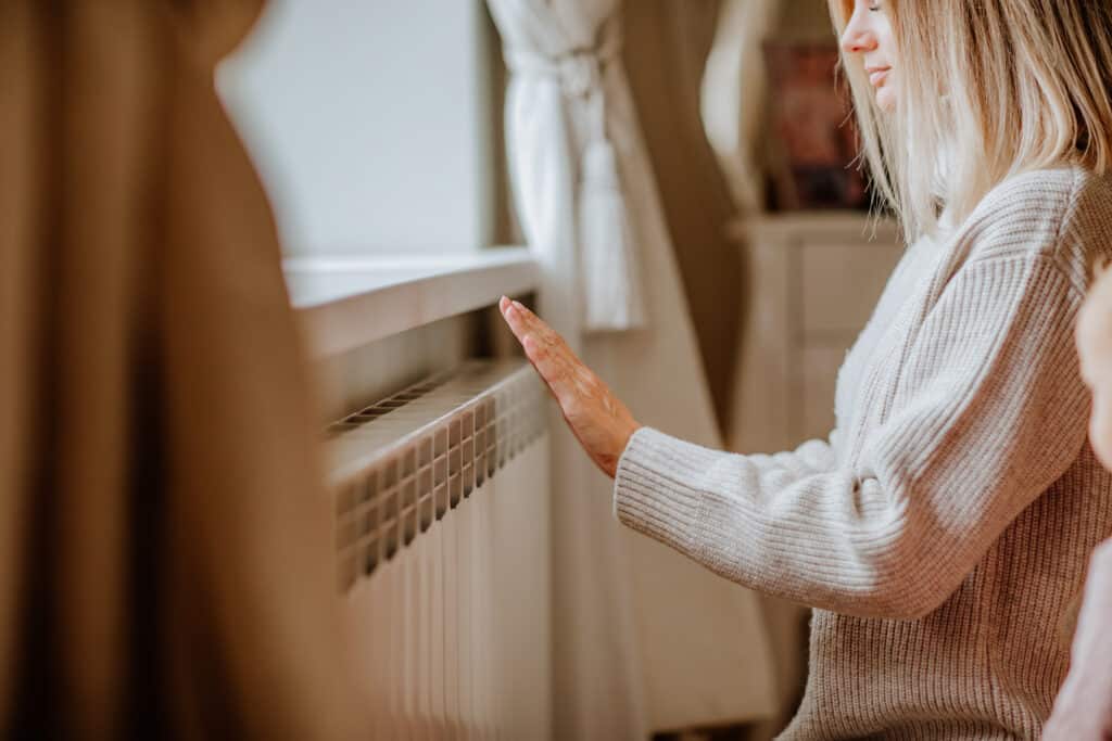 Femme en pull clair vérifiant la chaleur d'un radiateur blanc avec sa main, dans un intérieur cosy.