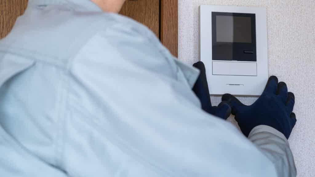 Technicien ganté en uniforme installant un interphone vidéo blanc sur un mur texturé près d'un cadre de porte en bois.