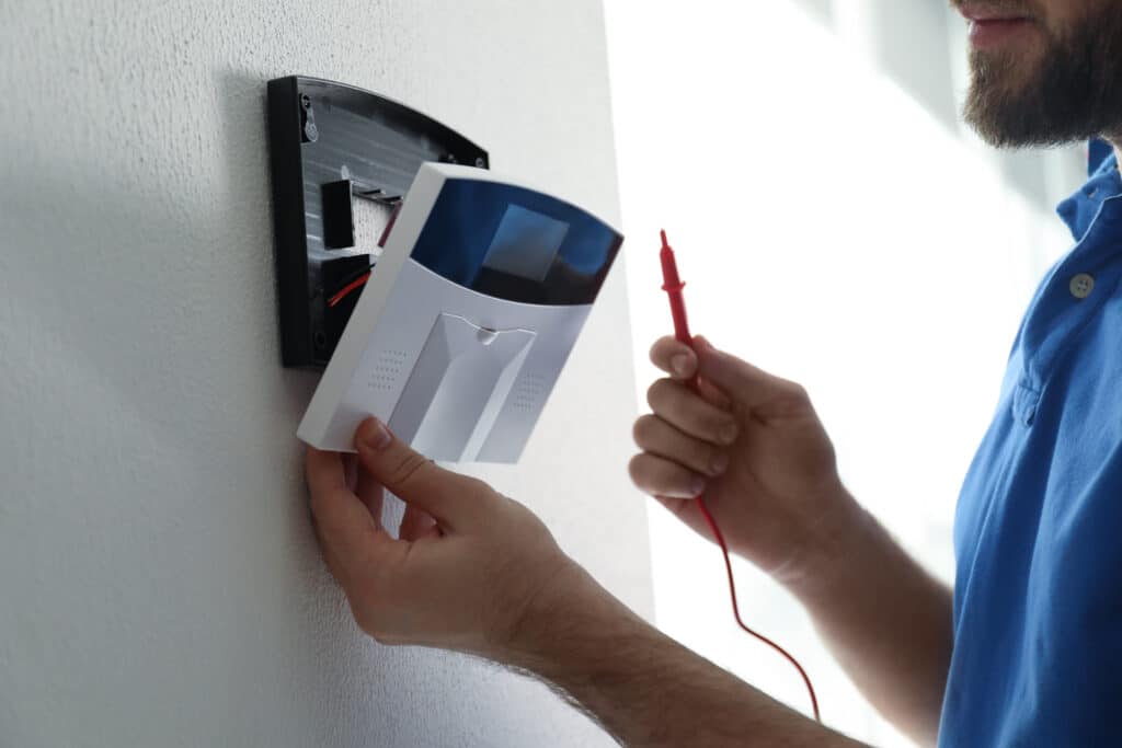 Homme barbu en polo bleu installant un système d'alarme blanc sur un mur texturé, tenant une sonde rouge pour vérification.