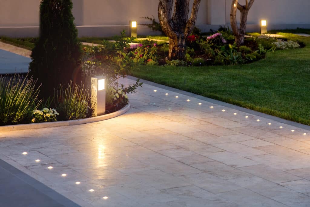 Allée pavée de jardin moderne illuminée la nuit par des spots encastrés et une borne lumineuse. Massifs de fleurs et pelouse luxuriante.