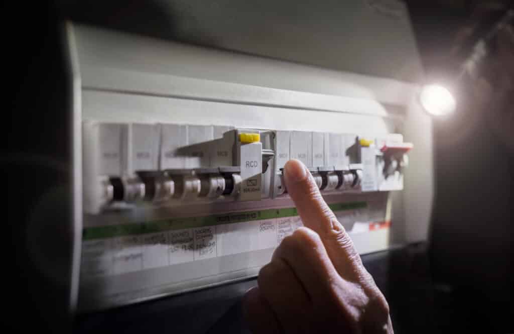 Main active un disjoncteur RCD sur un tableau électrique, éclairé par une lampe. Labels MCB, 80A 30mA et circuits (prises, lumières) visibles. Gestion coupure.