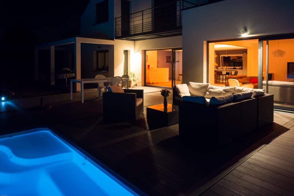 Villa contemporaine la nuit, patio en bois, piscine bleue lumineuse, salon de jardin et intérieur chaudement éclairé.